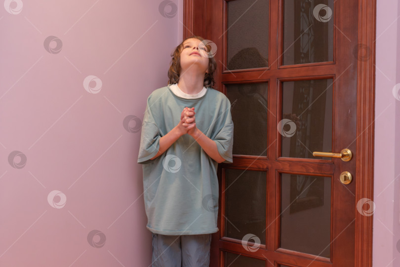 Скачать Маленький мальчик стоит в помещении, сложив руки, и смотрит вверх. Ребенок в углу рядом с деревянной дверью фотосток Ozero