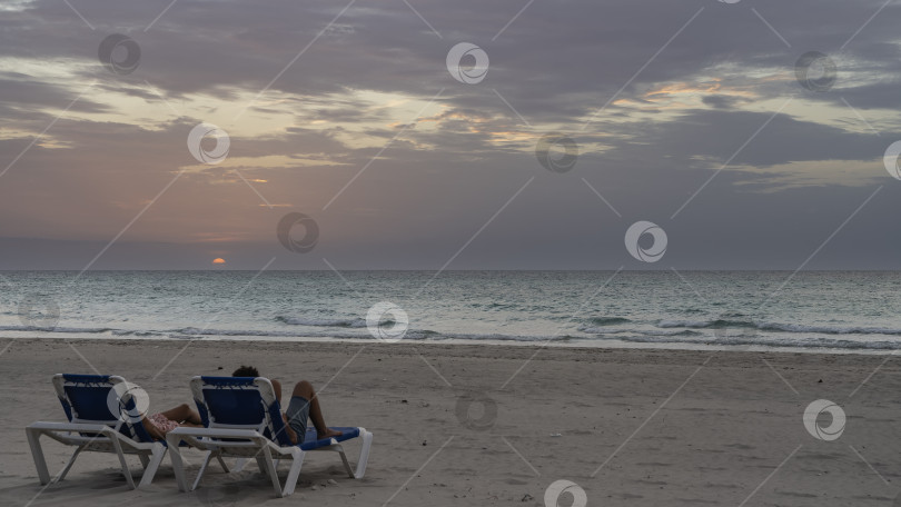 Скачать Два человека лежат в шезлонгах на песчаном пляже и наблюдают за закатом. фотосток Ozero