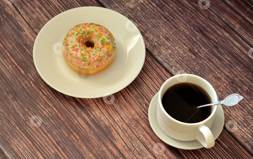 Скачать Свежий пончик в ванильной глазури с топпингом на тарелке и чашка черного кофе на деревянном столе. фотосток Ozero