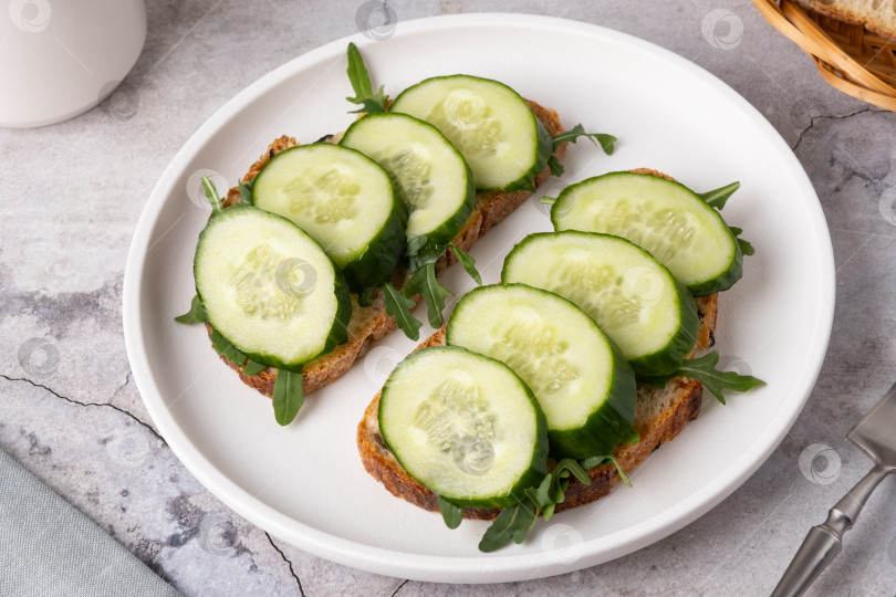Скачать Ломтики огурца на поджаренном хлебе с рукколой на простой тарелке в кухонной обстановке фотосток Ozero