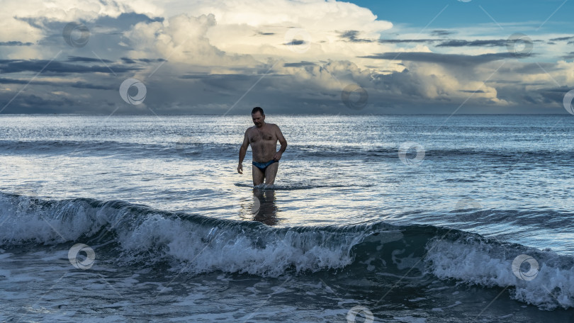 Скачать Раннее утро. Рассвет над океаном. Мужчина в плавках стоит в пенящихся волнах. фотосток Ozero