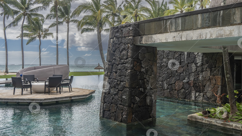 Скачать Открытый тропический бассейн. На деревянной террасе над водой установлены стулья и стол. фотосток Ozero