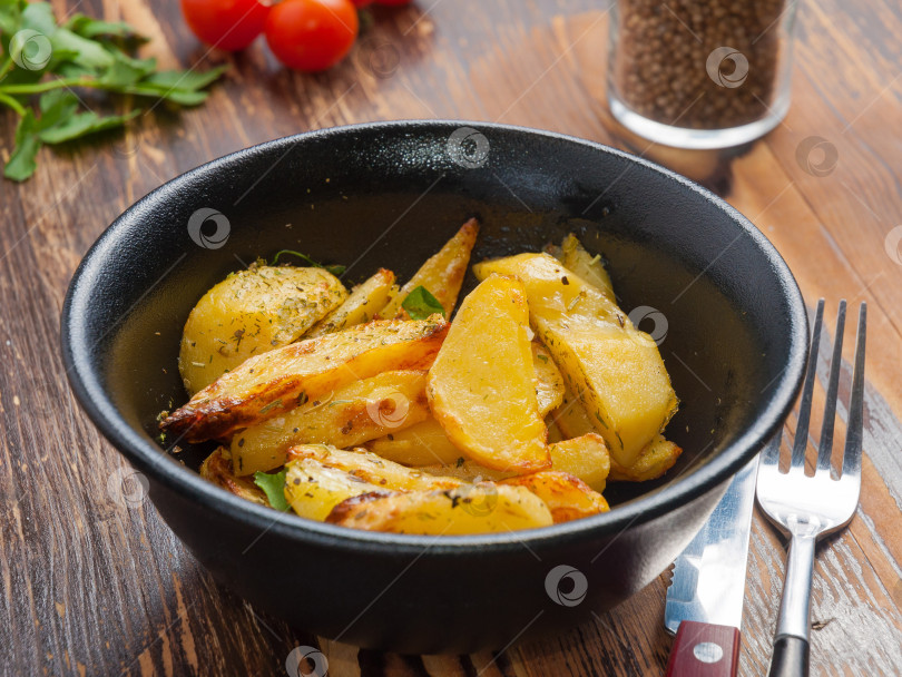 Скачать ломтики картофеля, запеченные с зеленью, в черной тарелке на деревянном столе фотосток Ozero