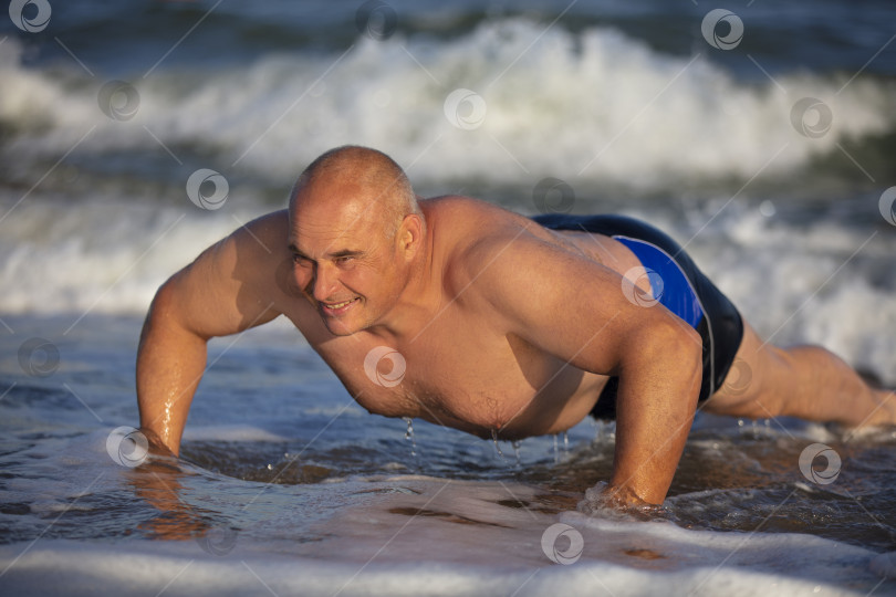 Скачать Пожилой мужчина делает зарядку на пляже. Мужчина отталкивается в воде. Активный отдых на пляже. фотосток Ozero