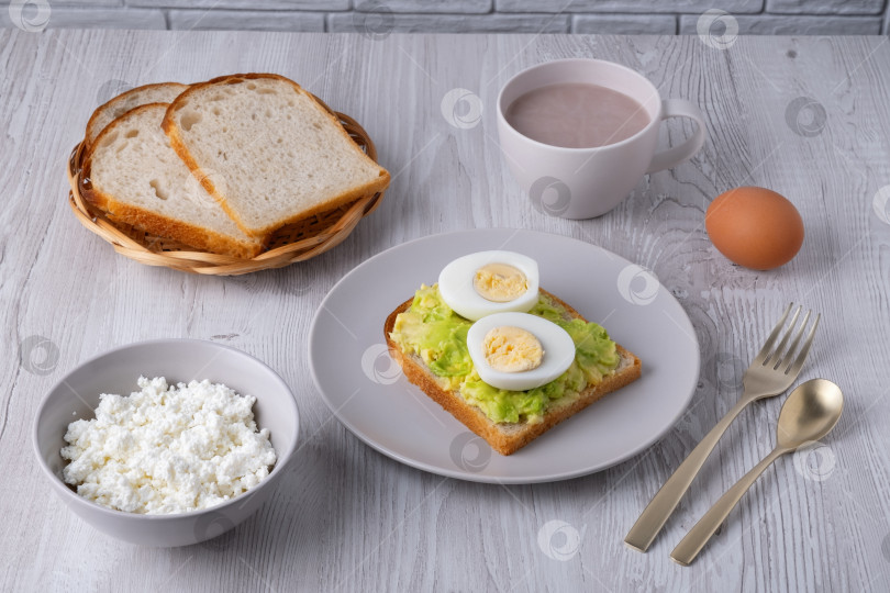 Скачать Полезный завтрак с тостами, вареными яйцами, творогом, хлебом и горячими напитками подается на деревянном столе в утреннем свете фотосток Ozero