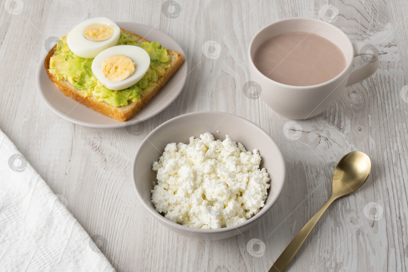 Скачать Полезный завтрак с тостами, вареными яйцами, творогом, хлебом и горячими напитками подается на деревянном столе в утреннем свете фотосток Ozero