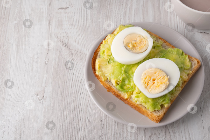 Скачать Тосты на завтрак с авокадо и вареными яйцами подаются на тарелке рядом с чашкой напитка в простой кухонной обстановке фотосток Ozero