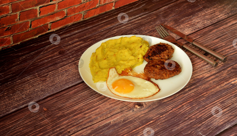 Скачать Тарелка со свежим картофельным пюре, яичницей-глазуньей и двумя мясными котлетами на деревянном столе, рядом столовые приборы. фотосток Ozero