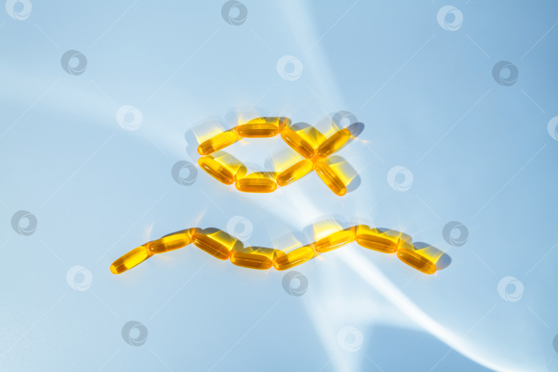 Скачать Желтые капсулы рыбьего жира омега-3 в форме рыбы с волнистой поверхностью, символизирующие натуральные добавки для укрепления здоровья и хорошего самочувствия, расположены на светло-голубом фоне фотосток Ozero
