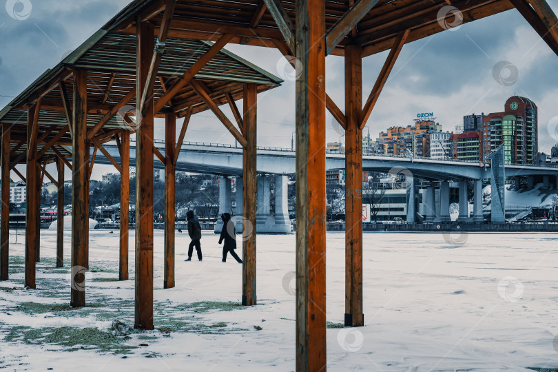 Скачать Зимний городской пейзаж с замерзшей рекой, деревянным навесом и Ворошиловским мостом на фоне. Январь 2026 год фотосток Ozero
