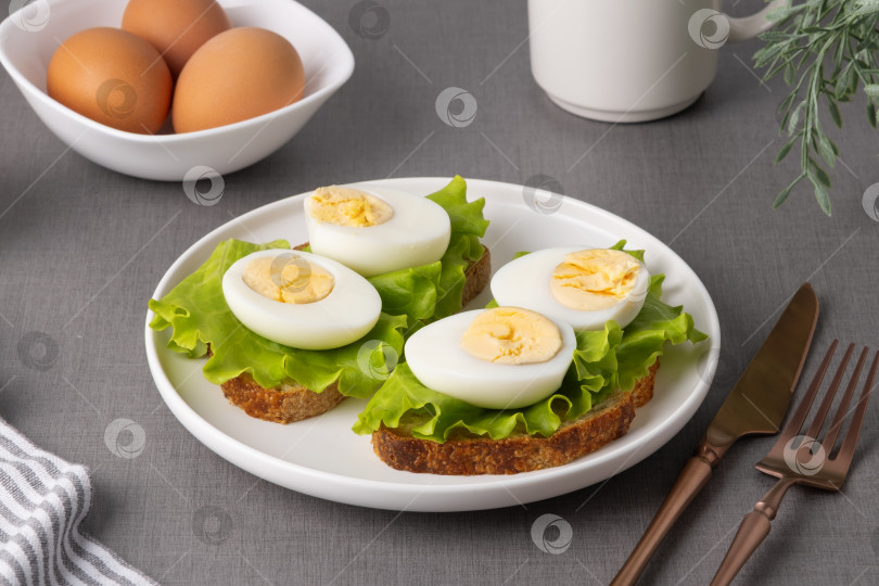 Скачать Сцена завтрака с вареными яйцами, листьями салата на хлебе, чаем и яйцами на сером столе с кухонными принадлежностями фотосток Ozero