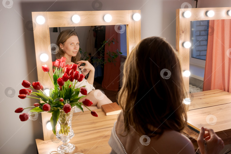 Скачать Женщина трогает ожерелье, глядя на отражение в зеркале с тюльпанами фотосток Ozero