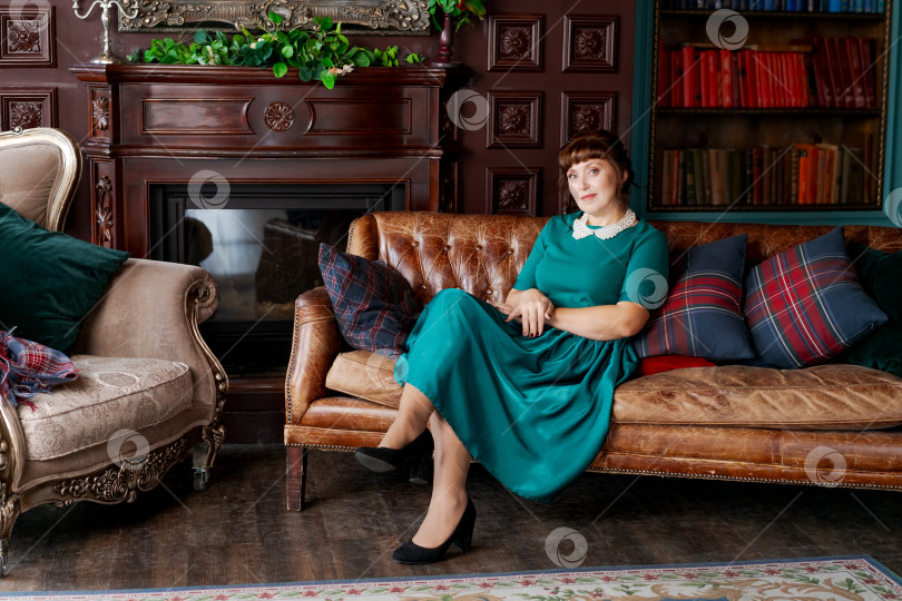 Скачать Элегантная зрелая женщина в зеленом платье сидит на кожаном диване у камина фотосток Ozero