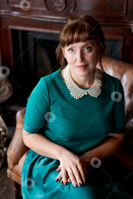 Скачать Уверенная в себе зрелая женщина в зеленом платье с жемчужным воротником сидит на диване фотосток Ozero