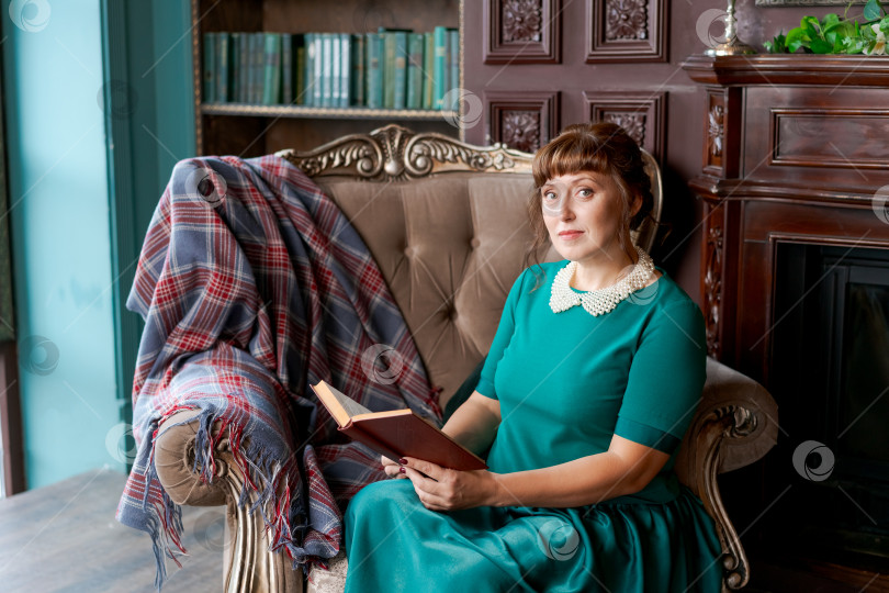 Скачать Уверенная в себе женщина с книгой сидит в элегантном кресле в Классической библиотеке фотосток Ozero