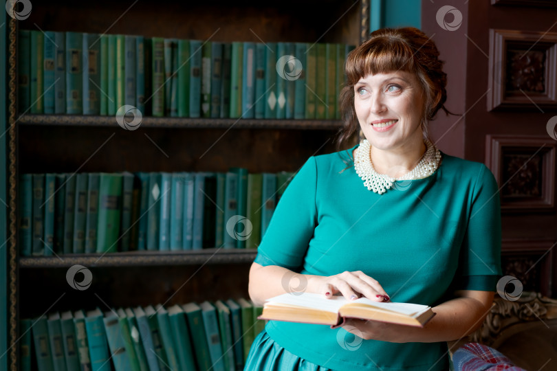 Скачать Радостная зрелая женщина с книгой, стоящая у книжной полки в Классической библиотеке фотосток Ozero