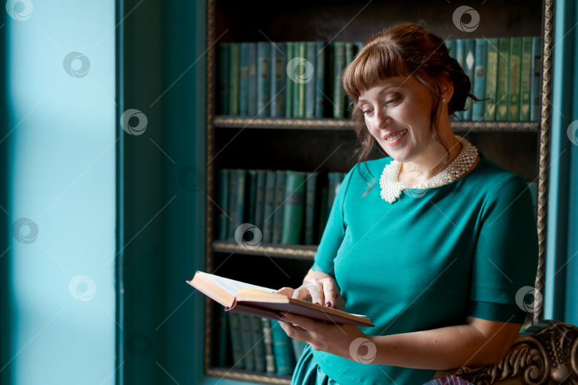 Скачать Счастливая зрелая женщина читает книгу и улыбается в классическом библиотечном интерьере фотосток Ozero