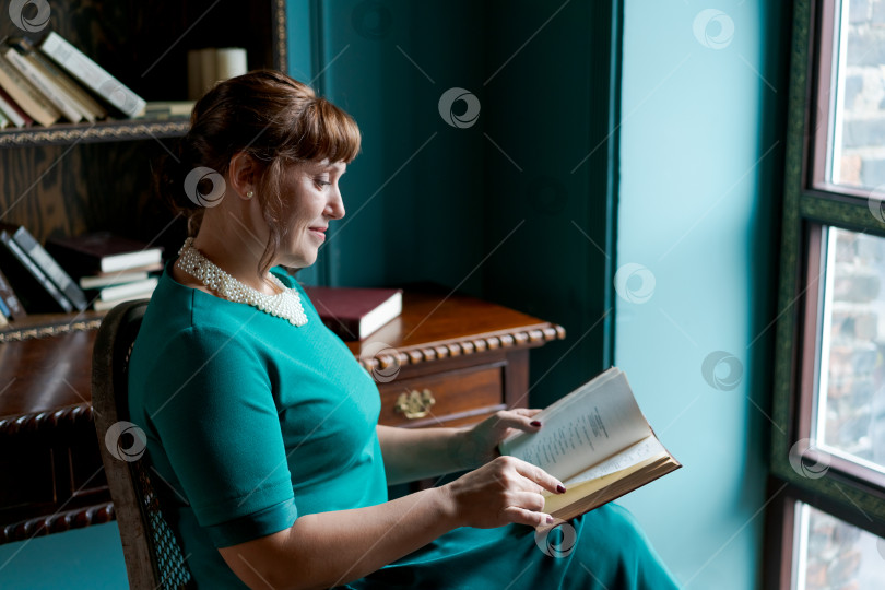 Скачать Элегантная женщина в профиль читает книгу у окна в классическом кабинете фотосток Ozero