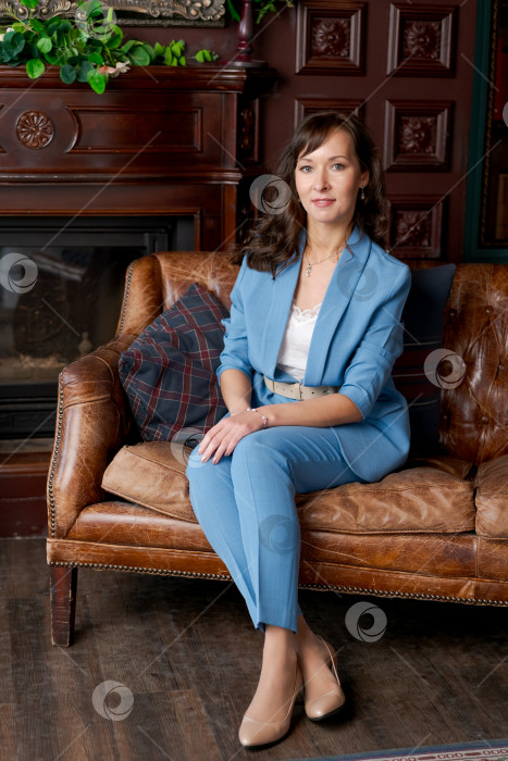 Скачать Уверенная в себе деловая женщина в синем костюме сидит на кожаном диване у камина фотосток Ozero