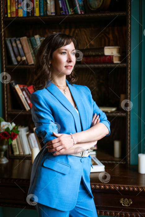 Скачать Уверенная в себе деловая женщина в синем костюме стоит у книжной полки в классическом офисе фотосток Ozero