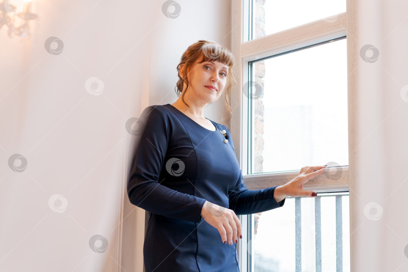 Скачать Уверенная в себе профессионалка в темном платье стоит у окна в светлом офисе фотосток Ozero