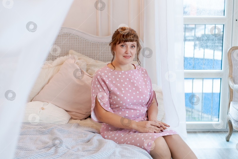 Скачать Прекрасная женщина в розовом платье в горошек сидит на кровати у окна фотосток Ozero