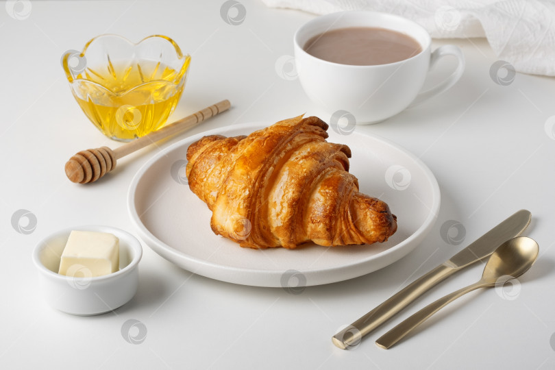 Скачать Завтрак состоит из круассана, меда, масла и горячего напитка и подается на белом столе с небольшим блюдечком и золотой ложечкой фотосток Ozero