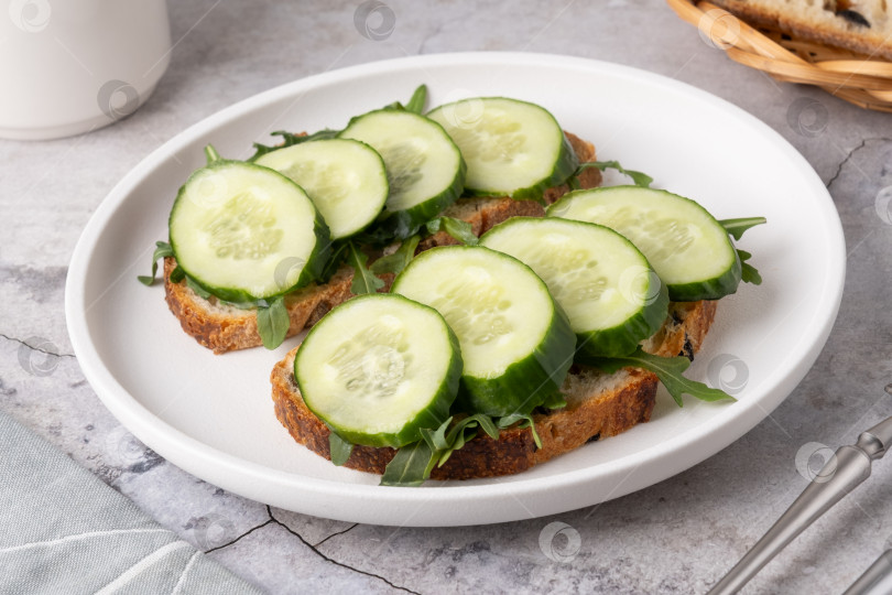 Скачать Ломтики огурца на поджаренном хлебе с рукколой на простой тарелке в кухонной обстановке фотосток Ozero