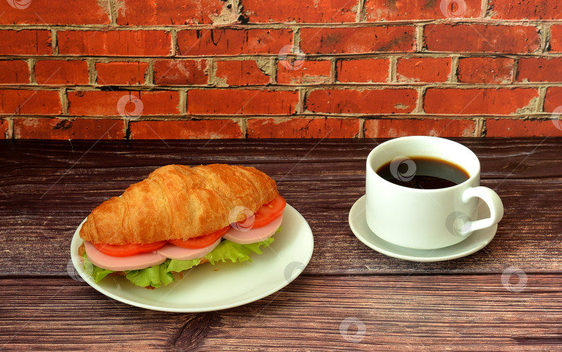 Скачать Бутерброд с круассаном, колбасой, спелыми помидорами и свежим листом салата-латука с чашечкой горячего черного кофе на деревянном столе. фотосток Ozero