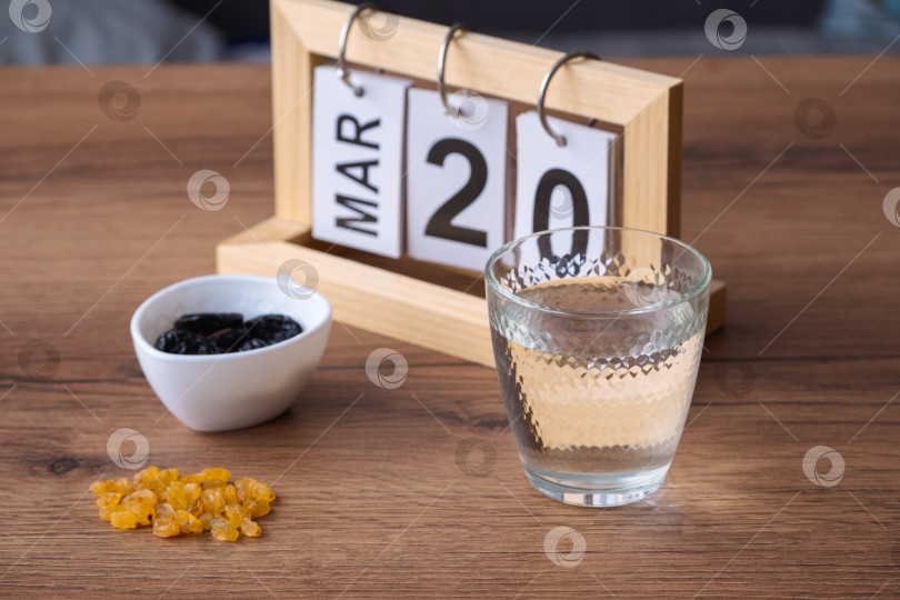 Скачать Простой натюрморт со стаканом воды, изюмом и черносливом, стоящими на деревянном столе, и деревянным календарем, показывающим 20 марта в честь окончания Рамадана. фотосток Ozero