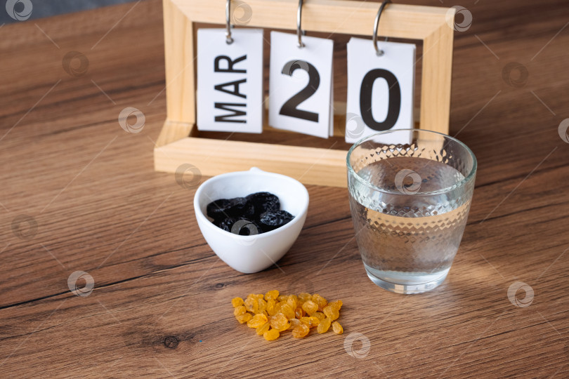 Скачать Простой натюрморт со стаканом воды, изюмом и черносливом, стоящими на деревянном столе, и деревянным календарем, показывающим 20 марта в честь окончания Рамадана. фотосток Ozero