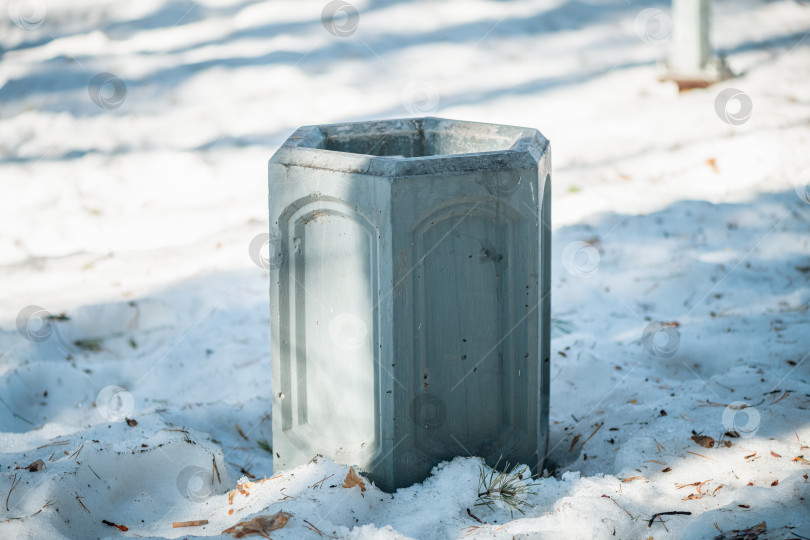 Скачать Шестиугольный бетонный мусорный бак, стоящий в парке, окруженный свежим белым снегом и разбросанными сосновыми иголками в ясный, холодный зимний день, символизирует чистоту и общественную полезность. фотосток Ozero
