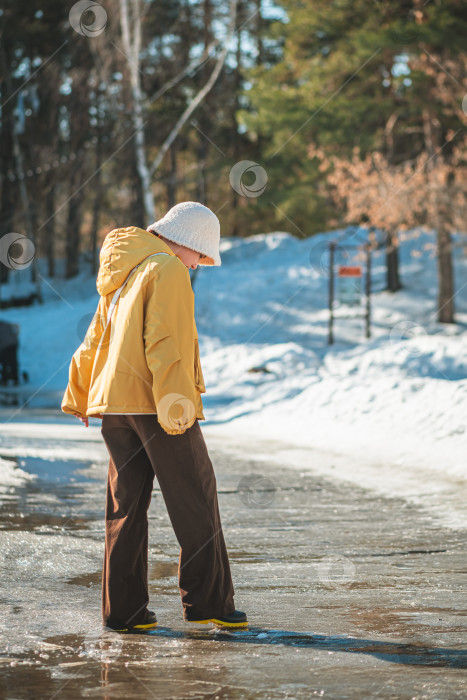 Скачать Девочка-подросток в желтой куртке и белой шапочке осторожно идет по обледенелой тропинке, устремив взгляд вниз, в окружении зимнего и весеннего лесного пейзажа и снежных заносов фотосток Ozero