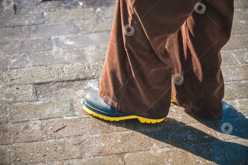 Скачать Человек стоит на выветренном кирпичном тротуаре, покрытом тающим льдом и слякотью, в широких коричневых вельветовых брюках и черных резиновых сапогах на ярко-желтой подошве, в весеннем уличном стиле фотосток Ozero