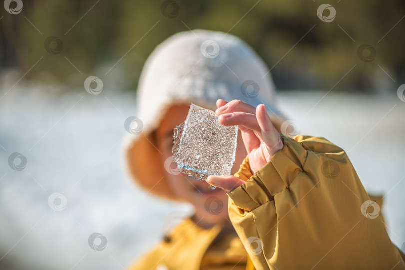 Скачать Человек в желтой куртке и кремовой шляпе, держащий голыми руками сверкающий кусочек кристаллического льда на размытом естественном фоне улицы, отражающий солнечный свет и ощущающий холод фотосток Ozero