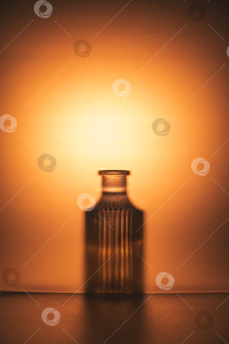 Скачать Стеклянный флакон создает эффектный силуэт на воздушном фоне оранжевого и янтарного цветов, создавая минималистичную композицию с безмятежной и теплой атмосферой. фотосток Ozero