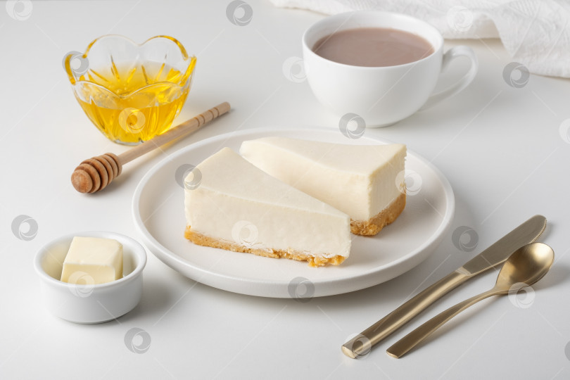 Скачать Ломтики чизкейка подаются с медом, сливочным маслом и чашкой горячего напитка на белом сервировочном столе. фотосток Ozero