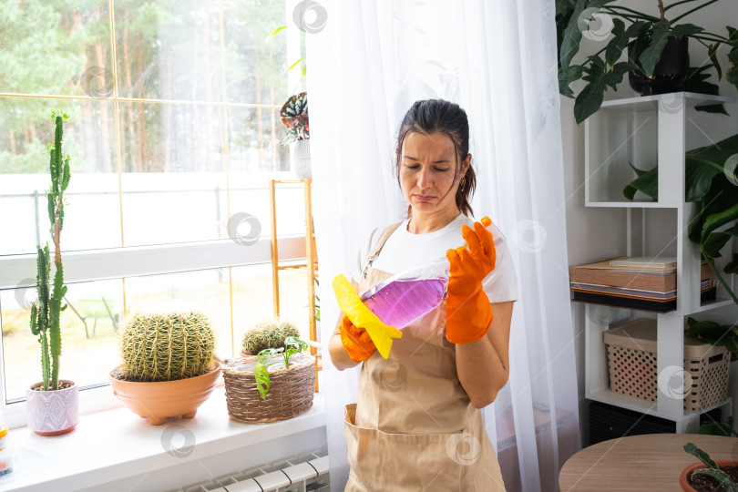 Скачать Женщина, недовольная работой по мытью окон, усталое выражение лица, перчатки и фартук, домашняя рутина, весеннее истощение, необходимость в поддержке клининговой компании, домашняя реклама фотосток Ozero