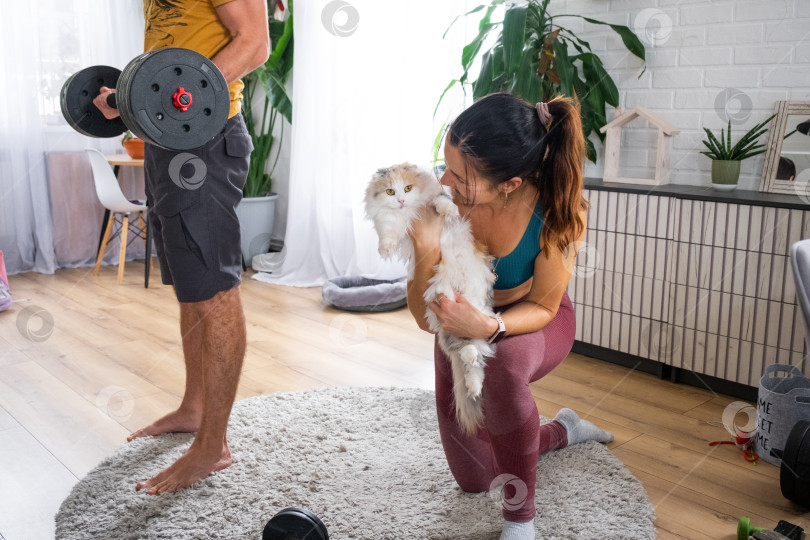 Скачать Женщина, мужчина и милая кошка сближаются после тренировки дома, уютный ковер, фитнес-образ жизни, семейное единение, радостная тренировка, любящая атмосфера. фотосток Ozero