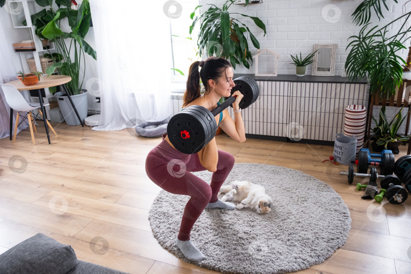 Скачать Молодая спортивная женщина тренируется со штангой в домашней обстановке, милый кот наблюдает за ней неподалеку, вдохновляющий фитнес-образ жизни, участие домашних животных, уютная атмосфера. фотосток Ozero