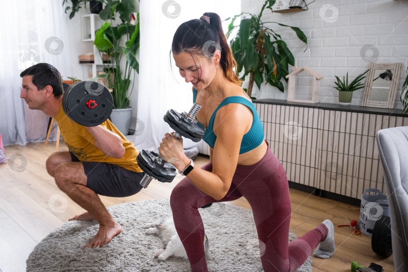 Скачать Женщина поднимает гантели с мужчиной в домашнем тренажерном зале, уютная комната с кошкой на коврике, семейный фитнес, совместные тренировки, отношения с домашними животными, современный образ жизни. фотосток Ozero