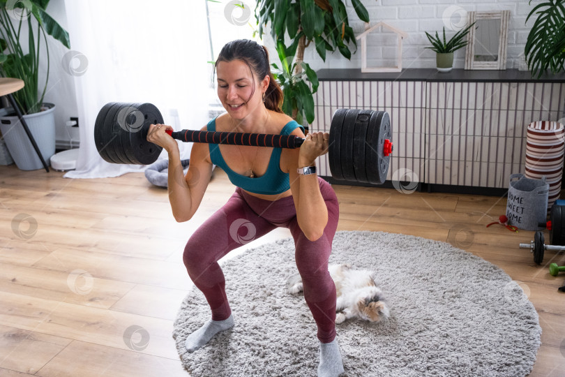 Скачать Улыбающаяся женщина, занимающаяся фитнесом дома с гантелями, игривый кот-компаньон на уютном коврике, здоровый распорядок дня, уход за домашними животными, современный интерьер. фотосток Ozero