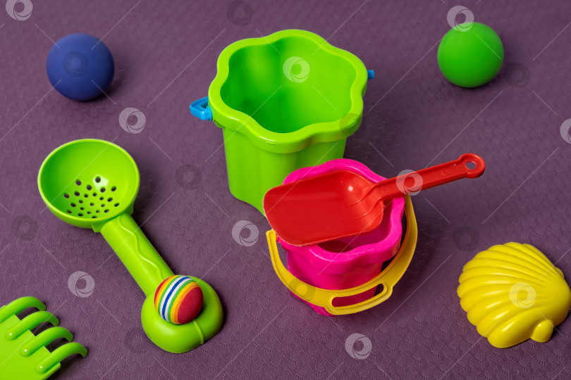 Скачать Яркие пляжные игрушки, разложенные на фиолетовой поверхности вместе с маленьким ведерком, совочком и шариками фотосток Ozero