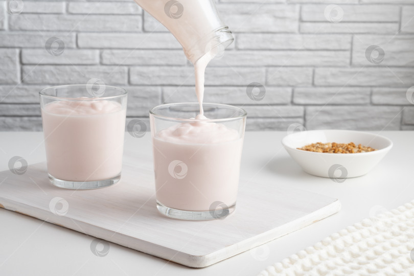 Скачать Заливка из домашнего кефира, пахты или йогурта с пробиотиками. Клубничный йогурт вытекает из стеклянной бутылки на светлом фоне. Холодный кисломолочный напиток с пробиотиками. Модные блюда и напитки фотосток Ozero