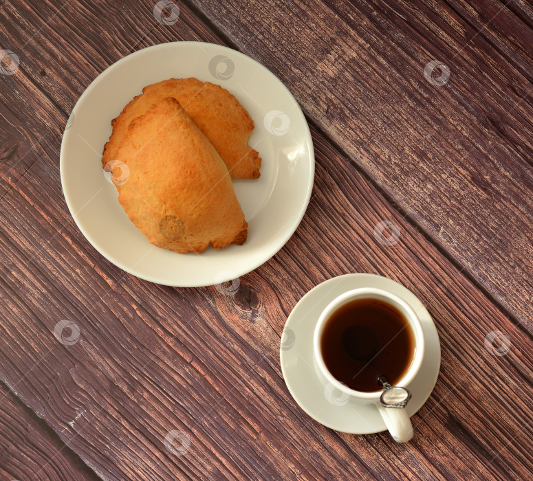 Скачать Чашка черного чая на блюдце с ложечкой и тарелка с двумя булочками из песочного теста с творожной начинкой на деревянном столе. фотосток Ozero