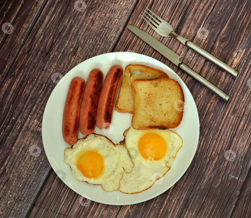 Скачать Плотный завтрак: два яичницы-глазуньи, сосиски и поджаренный хлеб на тарелке на деревянном столе. фотосток Ozero