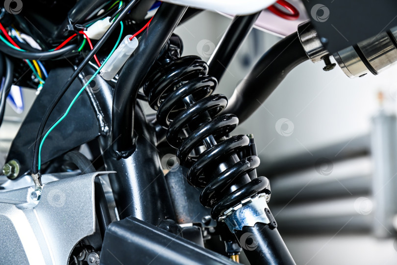 Скачать Крупный план заднего амортизатора мотоцикла с черной спиральной пружиной, металлической рамой и деталями электропроводки в гараже-мастерской без людей. фотосток Ozero
