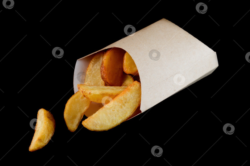 Скачать Хрустящая золотистая картофельная долька в кармашке из крафт-бумаги на черном фоне, вид сбоку крупным планом, с возможностью копирования, вкусная пикантная американская закуска из фаст-фуда фотосток Ozero