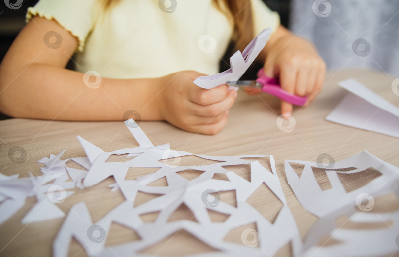 Скачать Ребенок создает дома за столом белые бумажные снежинки. Ребенок вырезает ножницами узоры на бумаге. Украшение для зимнего праздника. фотосток Ozero