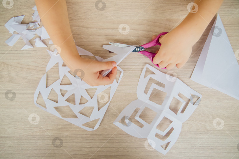 Скачать Ребенок создает дома за столом белые бумажные снежинки. Ребенок вырезает ножницами узоры на бумаге. Украшение для зимнего праздника. фотосток Ozero